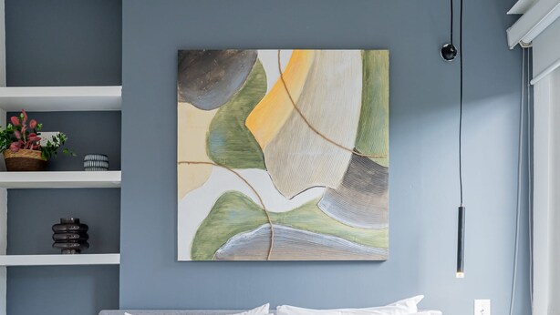 Un dormitorio con cama, almohadas blancas, un cuadro abstracto colgado en la pared y una mesita de noche con lámpara.