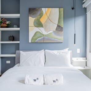 Un dormitorio con cama, almohadas blancas, un cuadro abstracto colgado en la pared y una mesita de noche con lámpara.