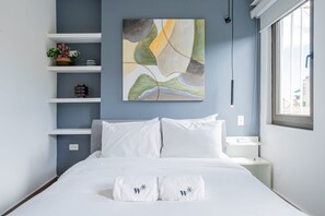 Una habitación de hotel moderna con cama, almohadas blancas, un cuadro abstracto colgado en la pared, una mesita de noche y una ventana con vistas.