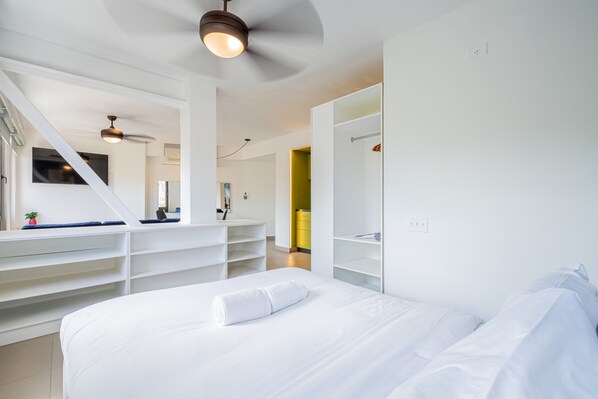 Una habitación de hotel moderna con una cama grande, estanterías integradas, un ventilador de techo y una paleta de colores neutros y luminosos.