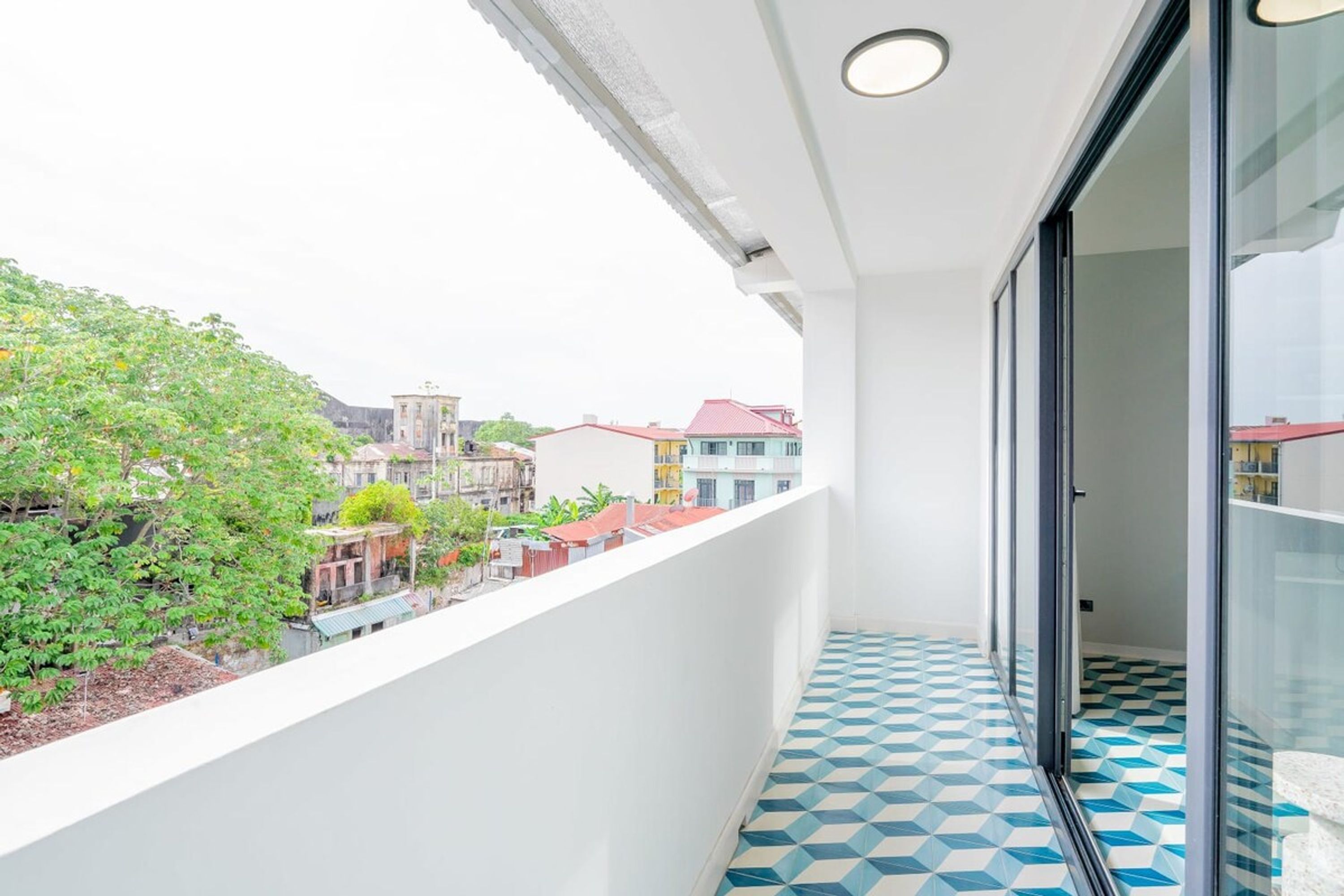 Un balcon doté d’un sol à motifs géométriques, d’une balustrade blanche et offrant une vue sur des immeubles et de la verdure.