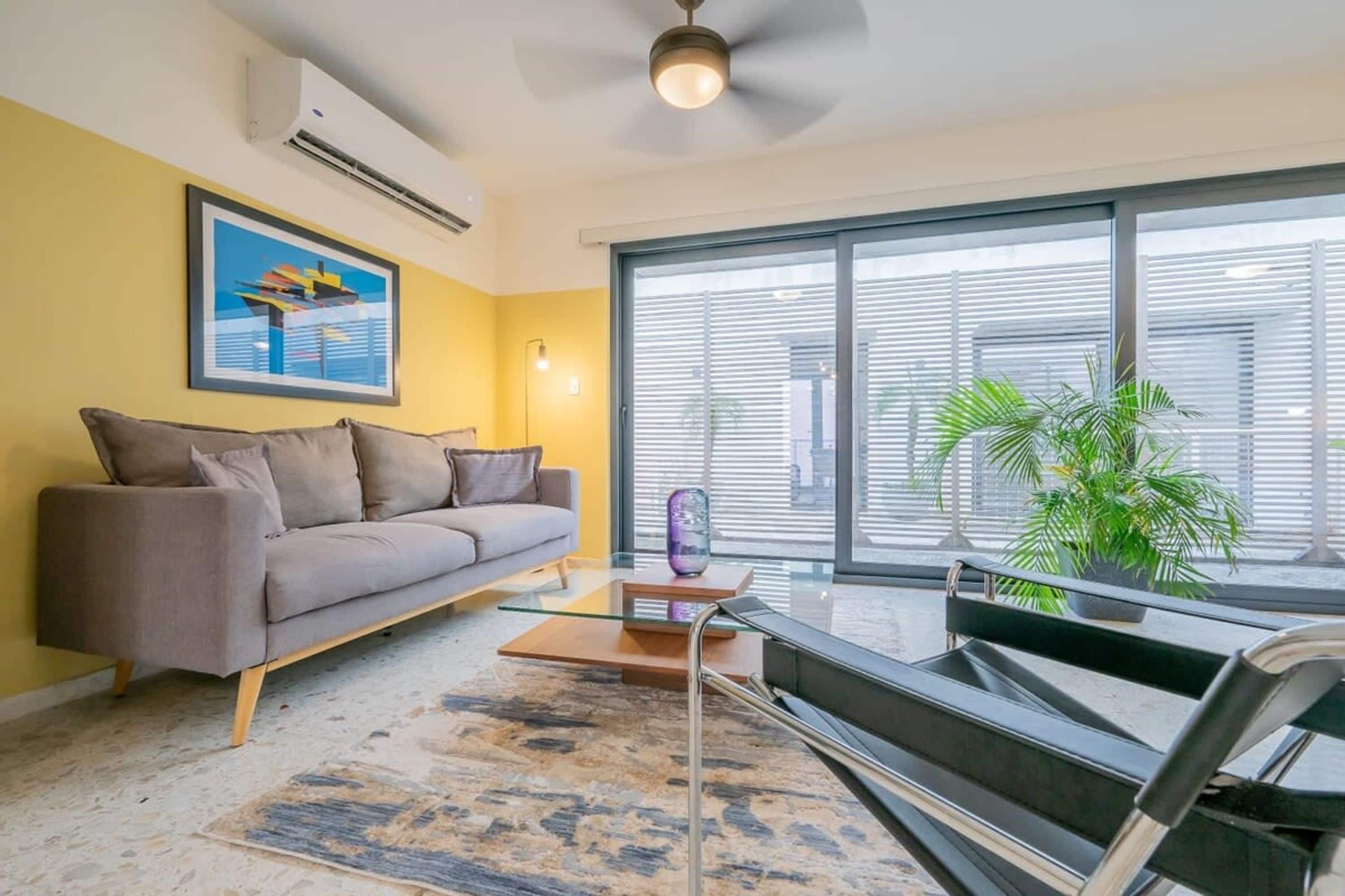 A modern living room with a gray sofa, a glass coffee table, and a large window with blinds.
