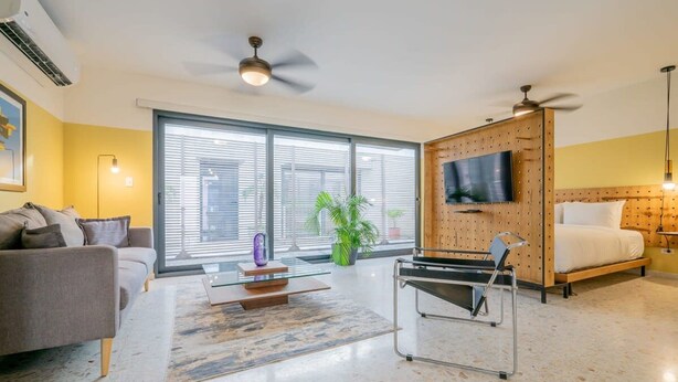 A modern living room with a sofa, a coffee table, a TV mounted on a wooden panel, and a bed with a headboard. - w Homey 1BR in Casco Antiguo (Panama City)