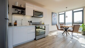 Une cuisine moderne avec des meubles blancs, un four encastré et un four à micro-ondes. On y trouve un coin repas avec une table et des chaises, ainsi qu’une grande fenêtre offrant une vue sur la ville.
