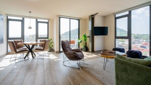Un vaste espace de vie comprenant une table à manger, des sièges confortables, une grande fenêtre offrant une vue sur la ville et un téléviseur à écran plat fixé au mur.