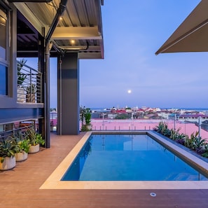 Ein moderner Pool im Freien mit einer Holzterrasse, Pflanzgefäßen und Blick auf eine Küstenstadt im Abendlicht.