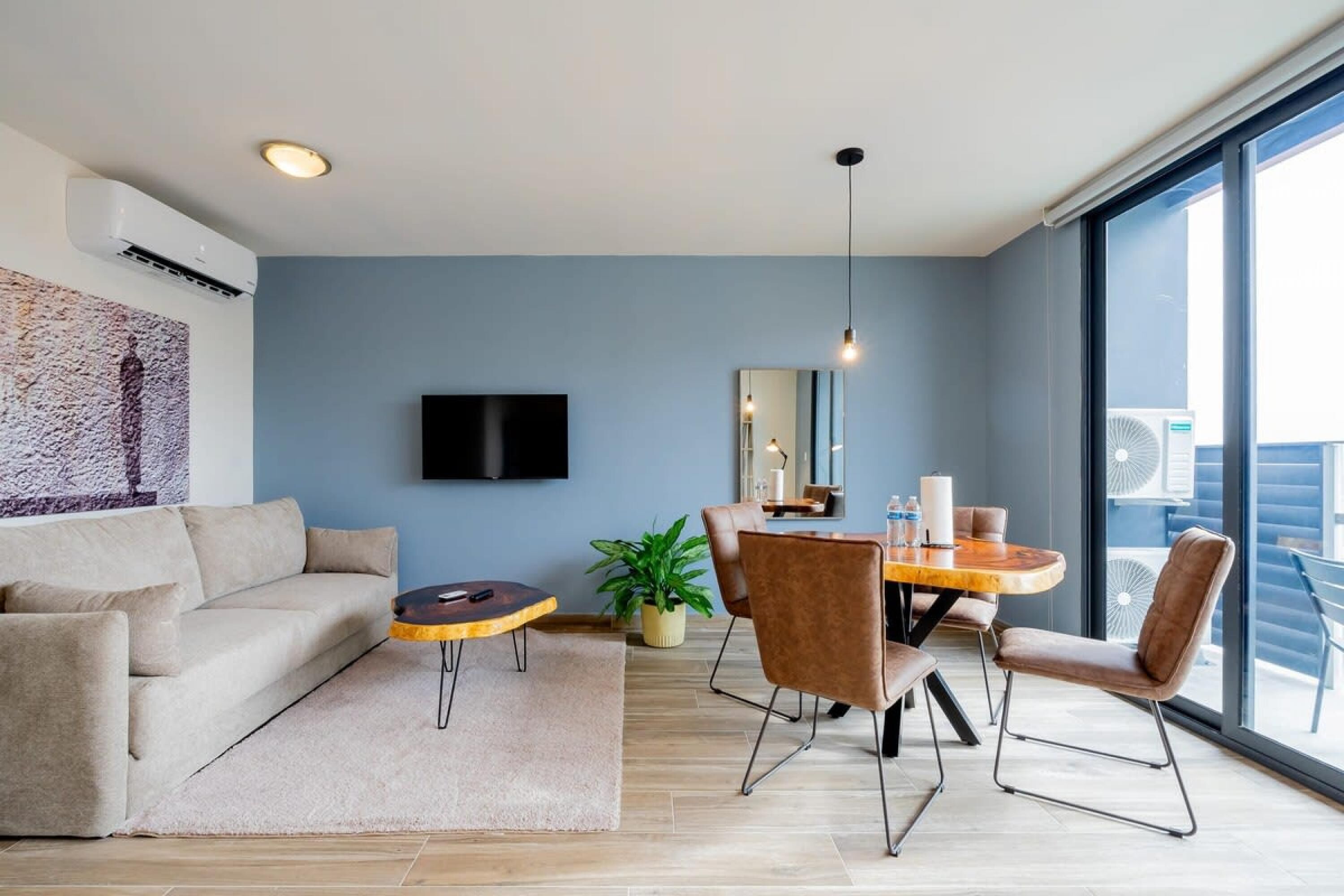 Una sala moderna con sofá, una mesa de centro, una mesa de comedor con sillas, un televisor montado en la pared y un amplio ventanal con vistas.