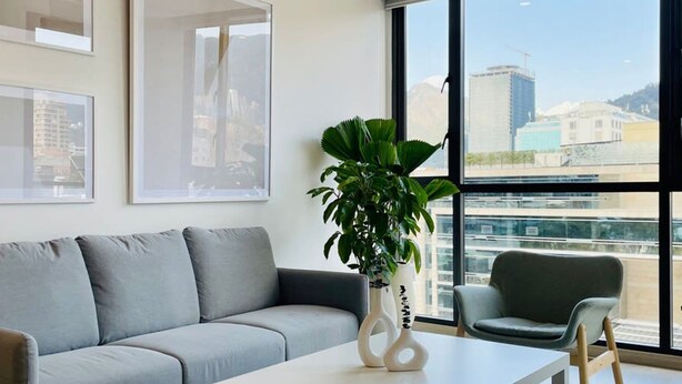 A modern living room with a grey sofa, a white coffee table, and a green plant. There are large windows with a view of buildings. - w Amazing 1BR With Balcony in Chico (Bogotá)