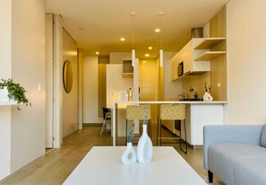 A modern living room with a white coffee table, two white vases, and a light blue sofa. There is a kitchen area with a white countertop and bar stools in the background. - w Amazing 1BR With Balcony in Chico (Bogotá)