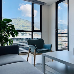 A modern living room with a sofa, a chair, and a table, large windows with a mountain view, and a potted plant. - w Amazing 1BR With Balcony in Chico (Bogotá)