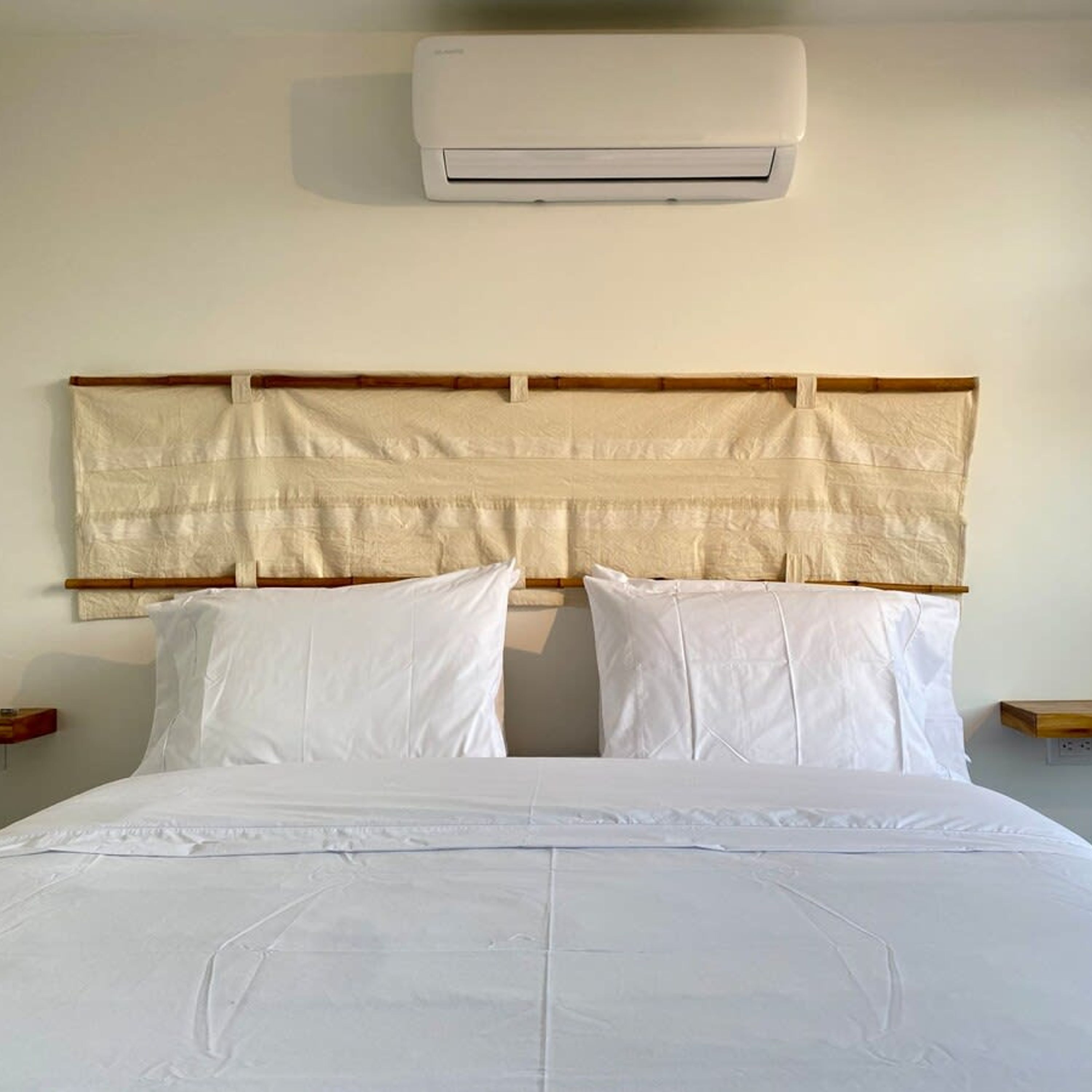 A hotel room with a bed, white bedding, a headboard with a woven design, and an air conditioning unit mounted on the wall.