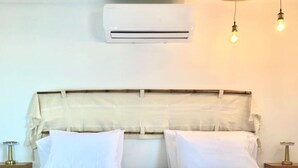Un lit bien fait, avec du linge de lit blanc, une tête de lit recouverte d’un tissu de couleur claire et deux tables de chevet avec des lampes. Un appareil de climatisation est fixé au mur, au-dessus du lit.