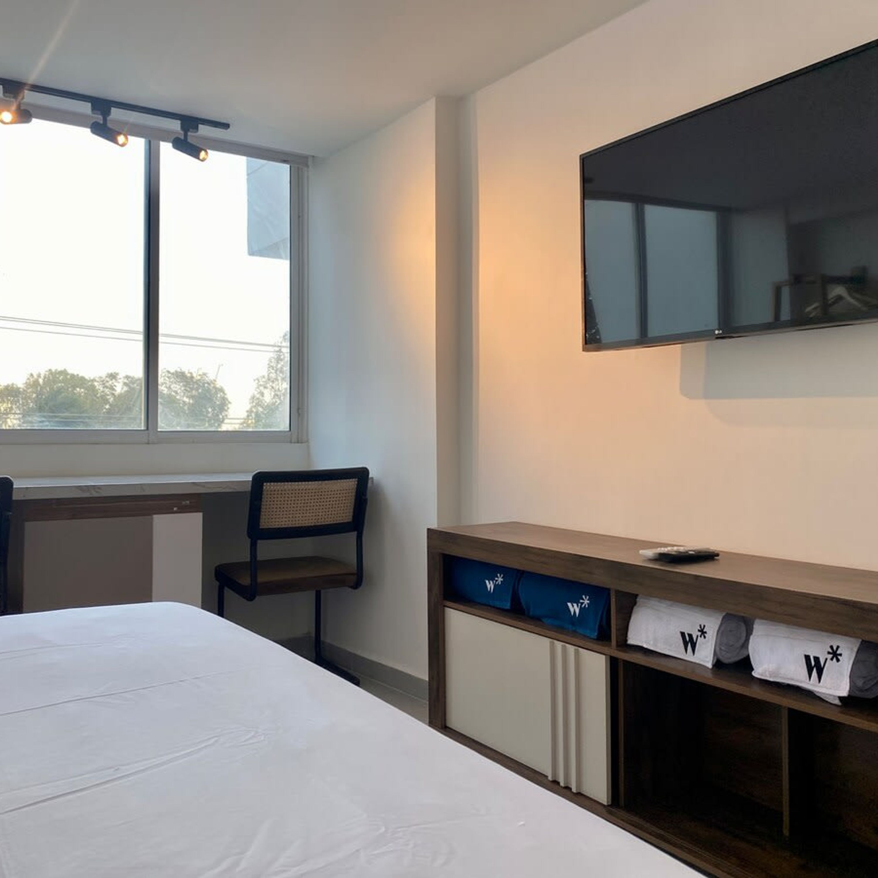 A hotel room with a bed, a chair, a desk, a TV mounted on the wall, and a window with a view of trees.