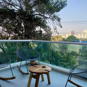 Un balcon avec deux fauteuils à bascule et une petite table en bois, d’où l’on peut admirer un paysage urbain avec ses bâtiments et sa verdure.