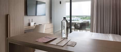 A modern hotel room with a wooden desk, a flat-screen TV, a large window with curtains, and a balcony with outdoor furniture.