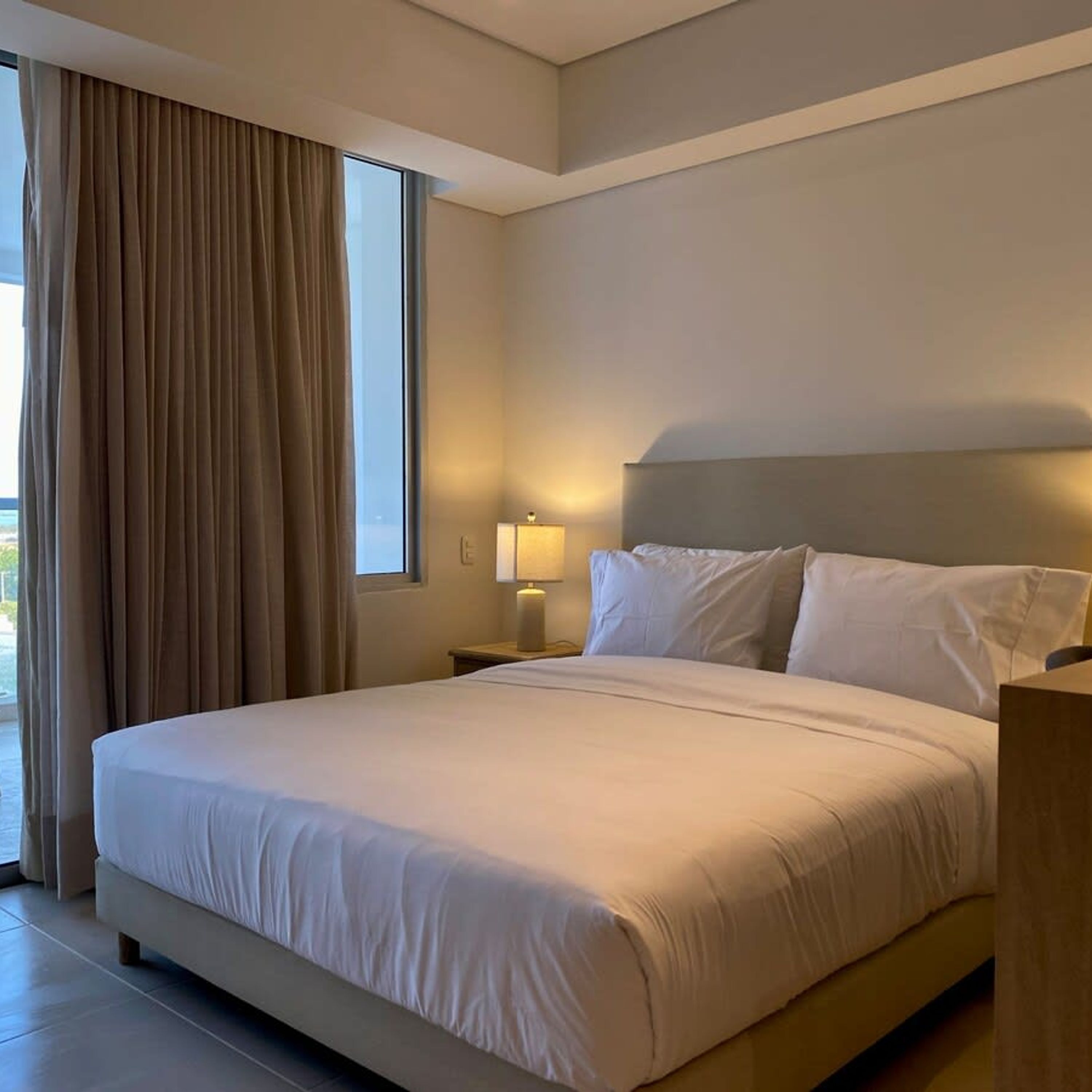 A hotel room with a large bed, a nightstand with a lamp, and a view through a window with curtains.