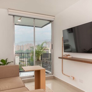 A modern apartment with a sliding glass door leading to a balcony, a flat-screen TV mounted on the wall, and a small table with books.