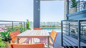 En balkon med et træbord og orange stole, med udsigt over en swimmingpool og høje bygninger.