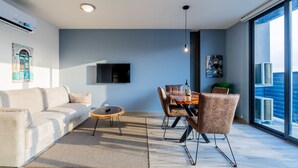 En moderne stue med sofa, sofabord, spisebord og stole. Der er et stort fjernsyn monteret på væggen, et stort vindue med udsigt og en aircondition-enhed på væggen.