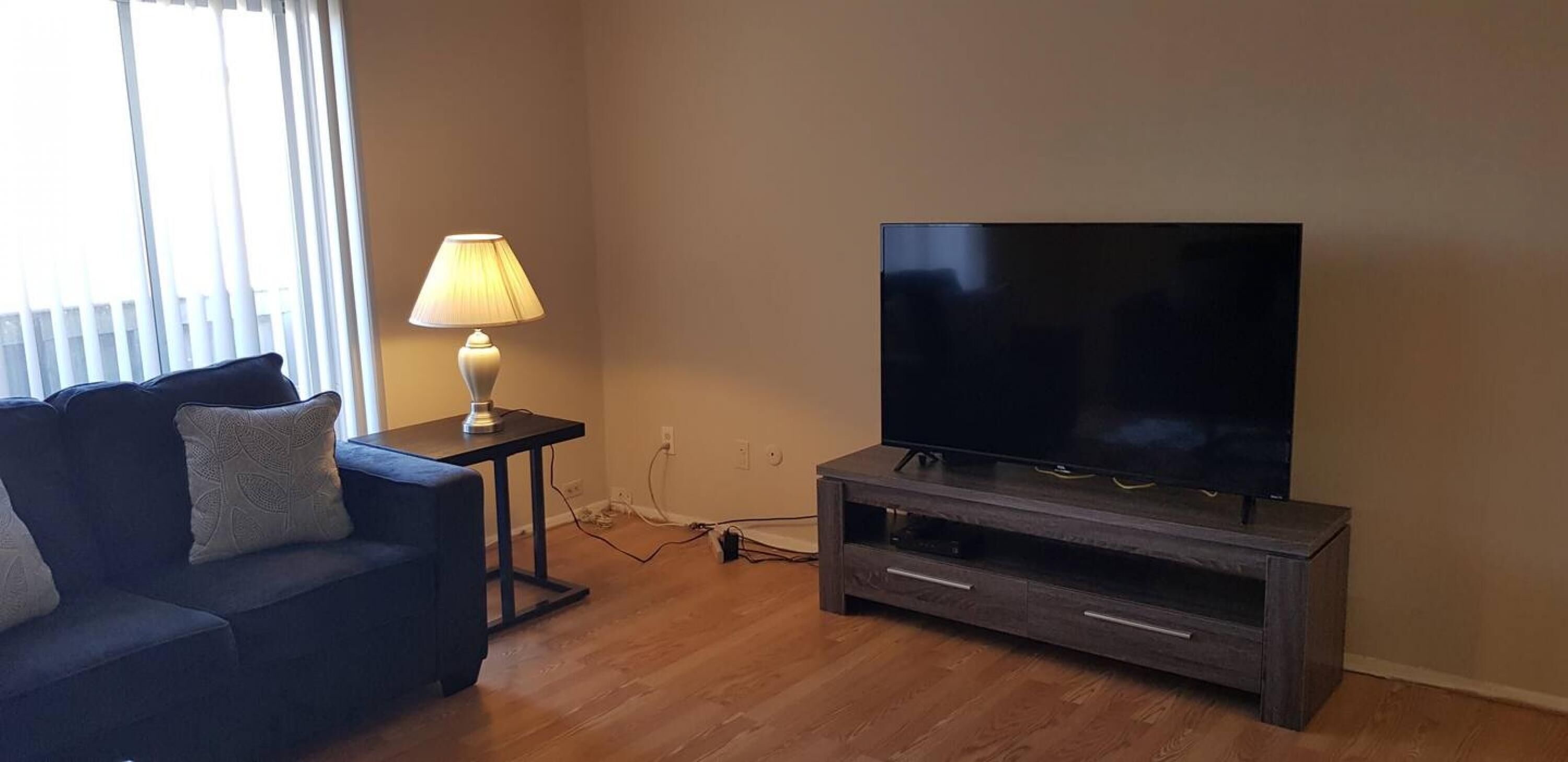 A living room with a dark wood TV stand, a flat-screen TV, a dark blue sofa with patterned cushions, and a table lamp.