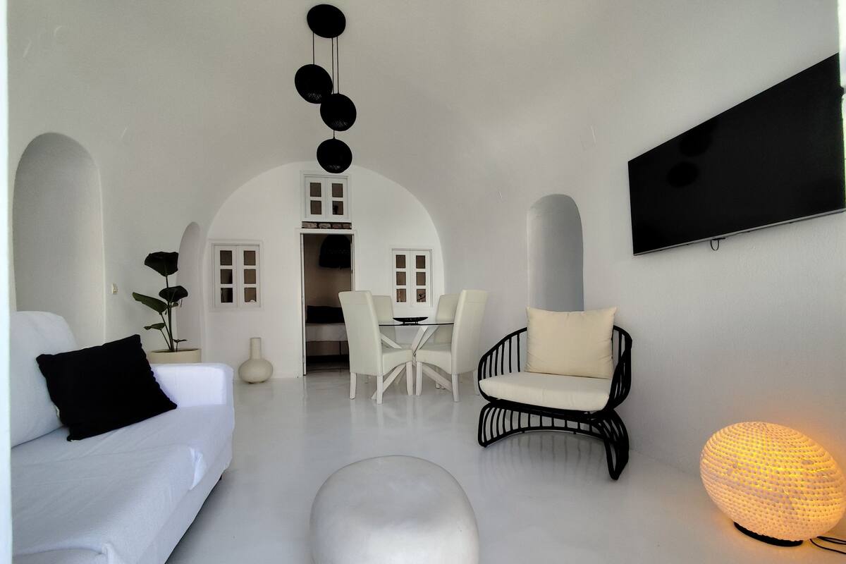 A modern, minimalist living room with a white sofa, a black armchair, a dining table with chairs, and a large TV mounted on the wall.