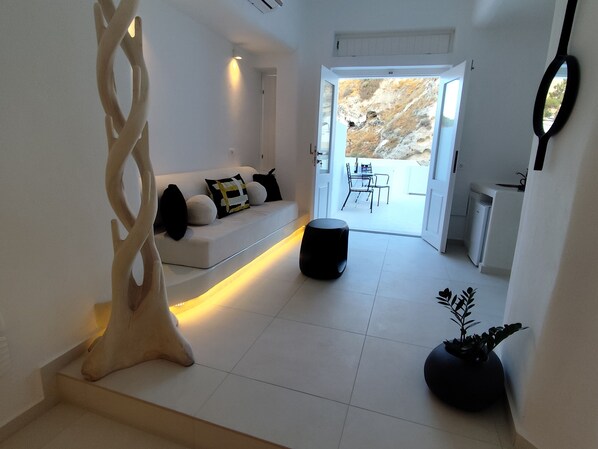 A modern living room with a white sofa, black and white cushions, a black ottoman, and a decorative wooden piece. A potted plant is on the floor, and a dining area is visible through an open door. - The Magic Luxury Cave Suites (Santorini)