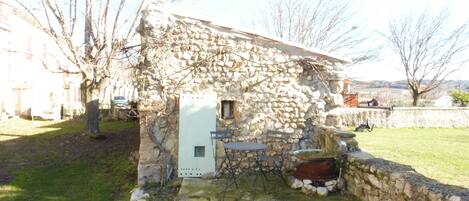 Un edificio de piedra con una ventana pequeña y una puerta, rodeado por un muro de piedra y un área con césped.