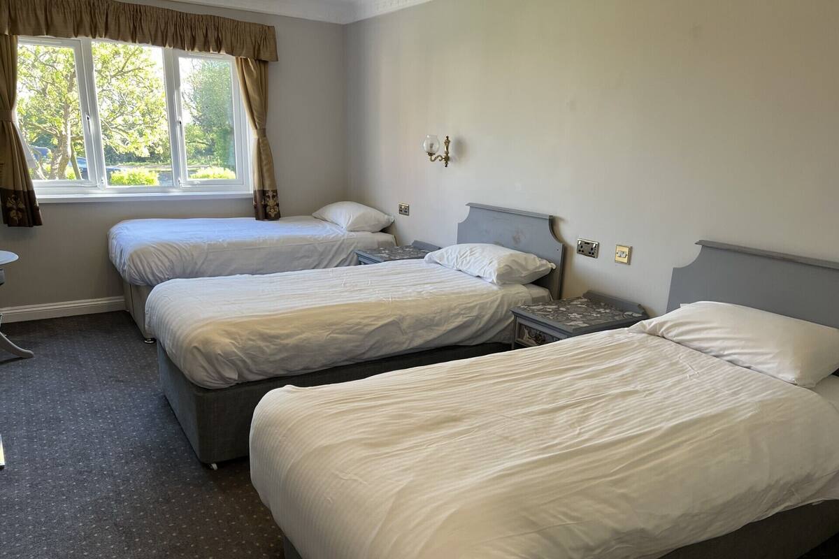 A hotel room with two single beds, a small table, a nightstand, a window with curtains, and a wall-mounted light fixture.