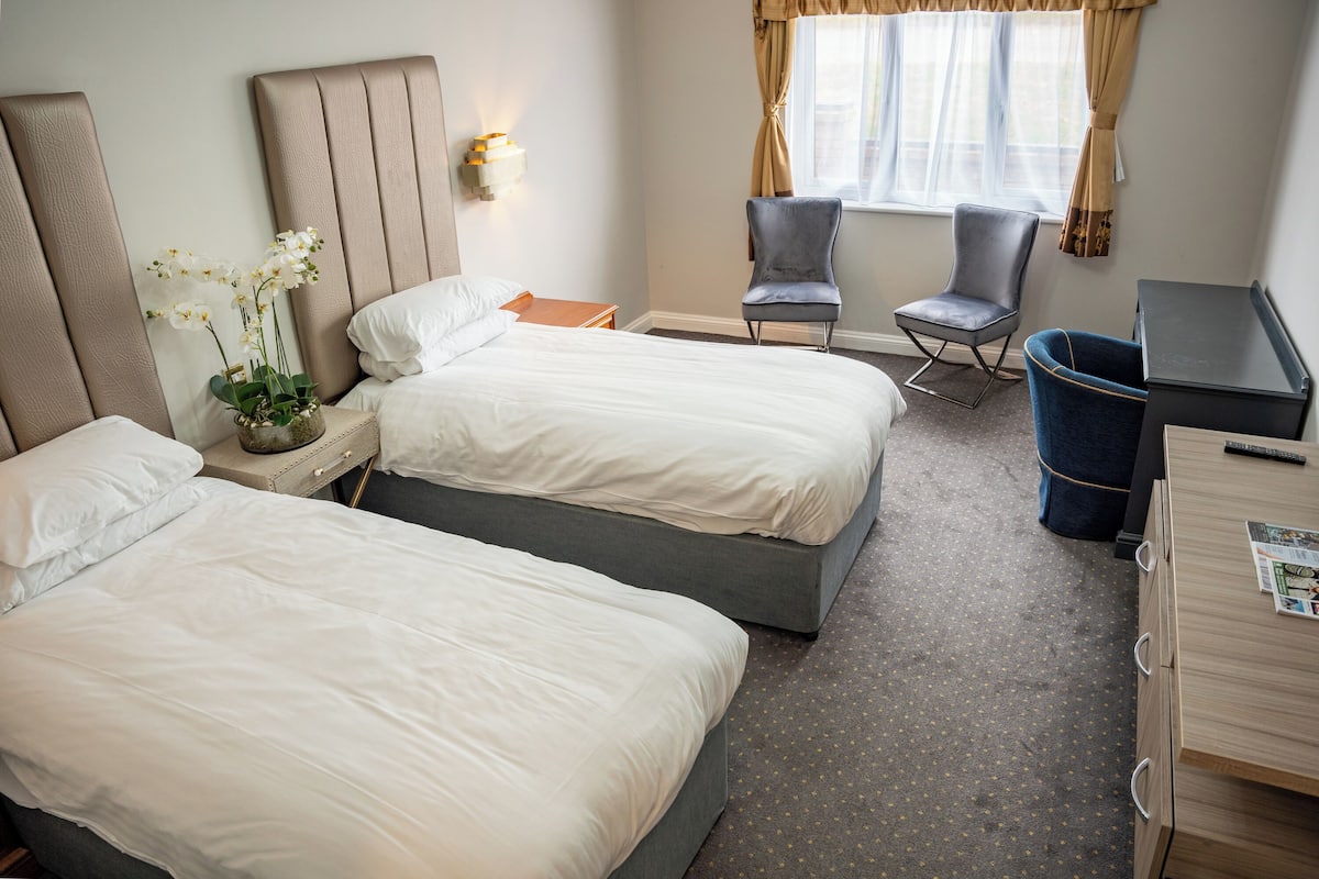 A hotel room with two beds, a desk, a chair, a trash bin, and a window with curtains.
