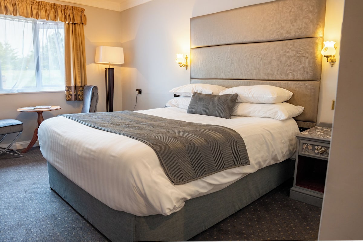 A neatly made bed with a grey blanket, a bedside table with a lamp, and a chair in a hotel room.