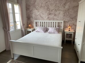 Ein Schlafzimmer mit einem weißen Bett, zwei Nachttischen mit Lampen, einem Kleiderschrank und einem Fenster mit Vorhängen.