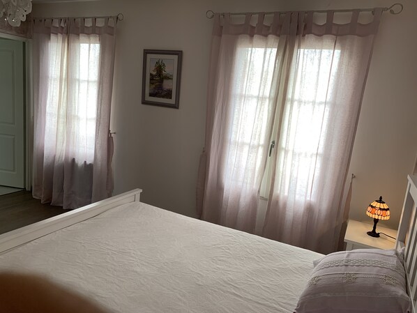 A bedroom with a bed, a nightstand with a lamp, and a window with sheer curtains.
