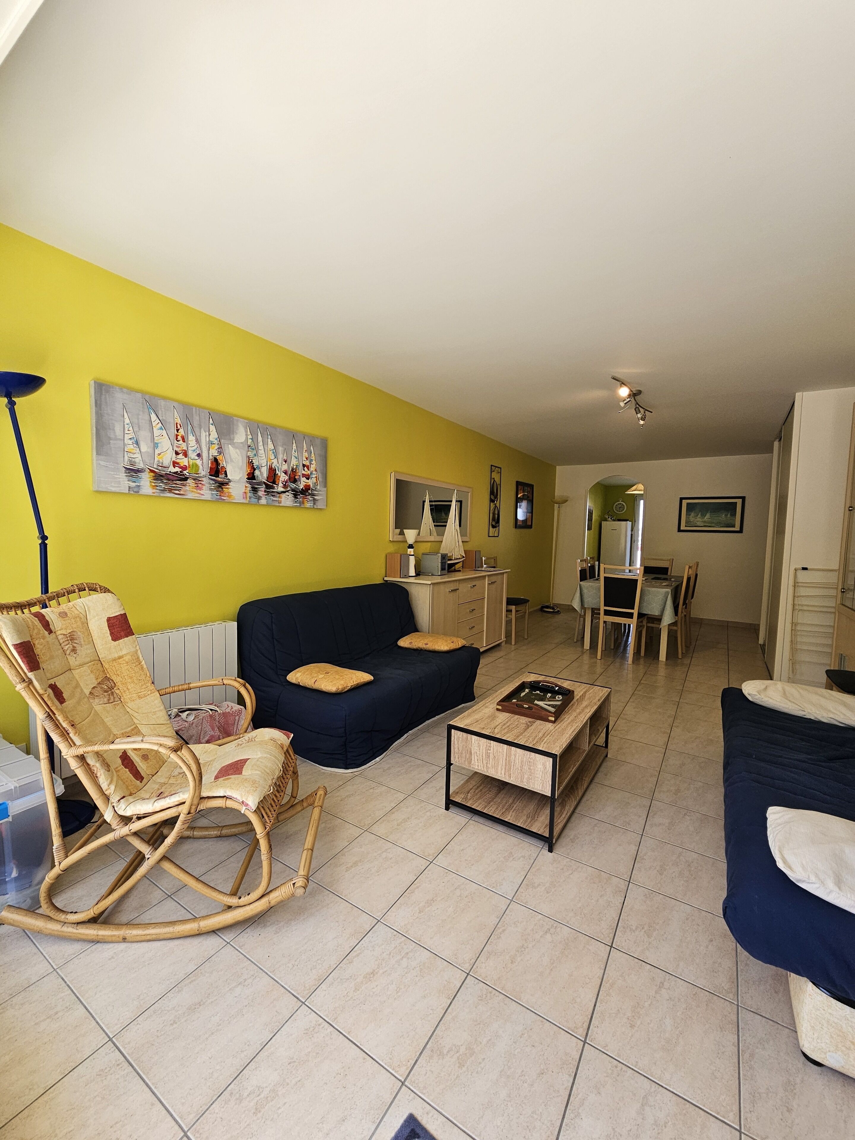 A living room with a yellow wall, a black sofa, a wooden coffee table, a wicker rocking chair, and a dining area with a table and chairs.