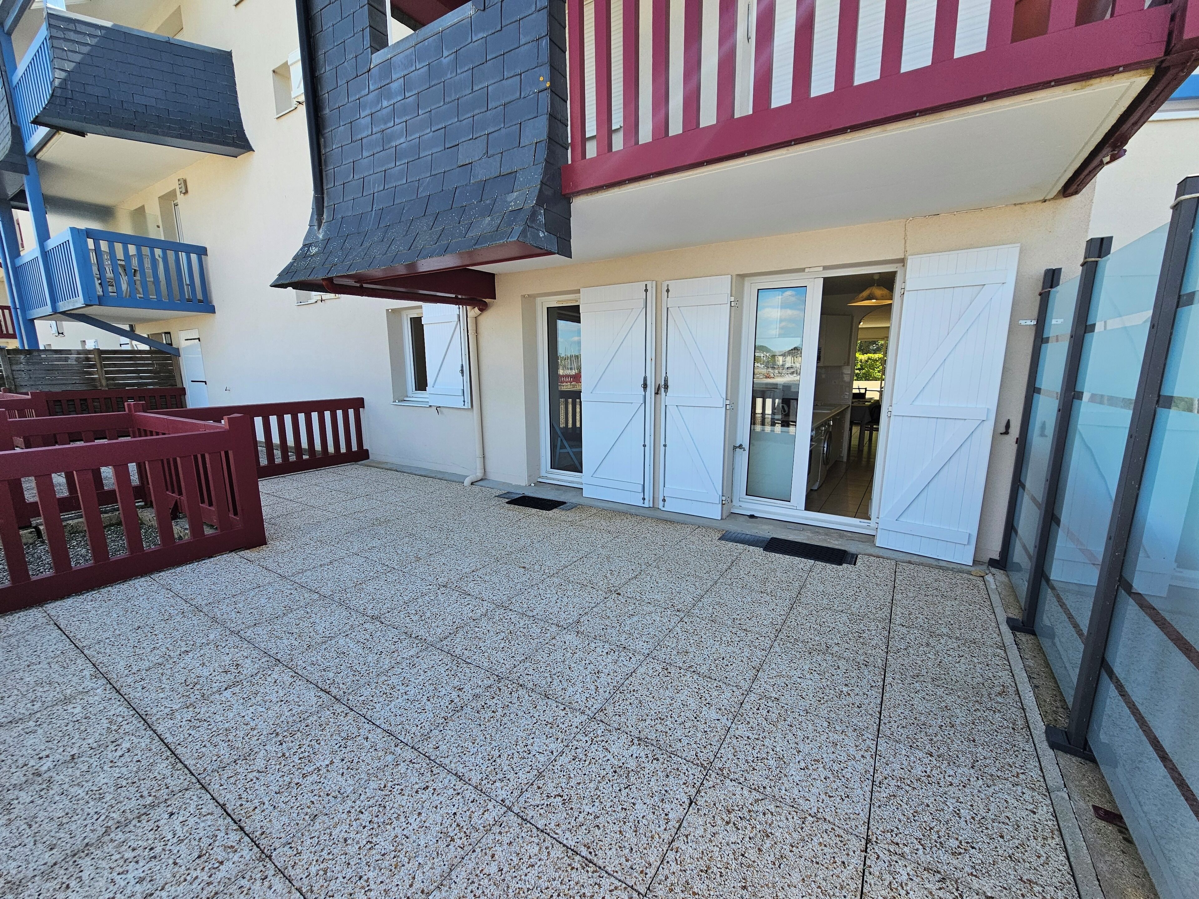 A courtyard with a tiled floor, a red fence, and a building with a red balcony and white doors.