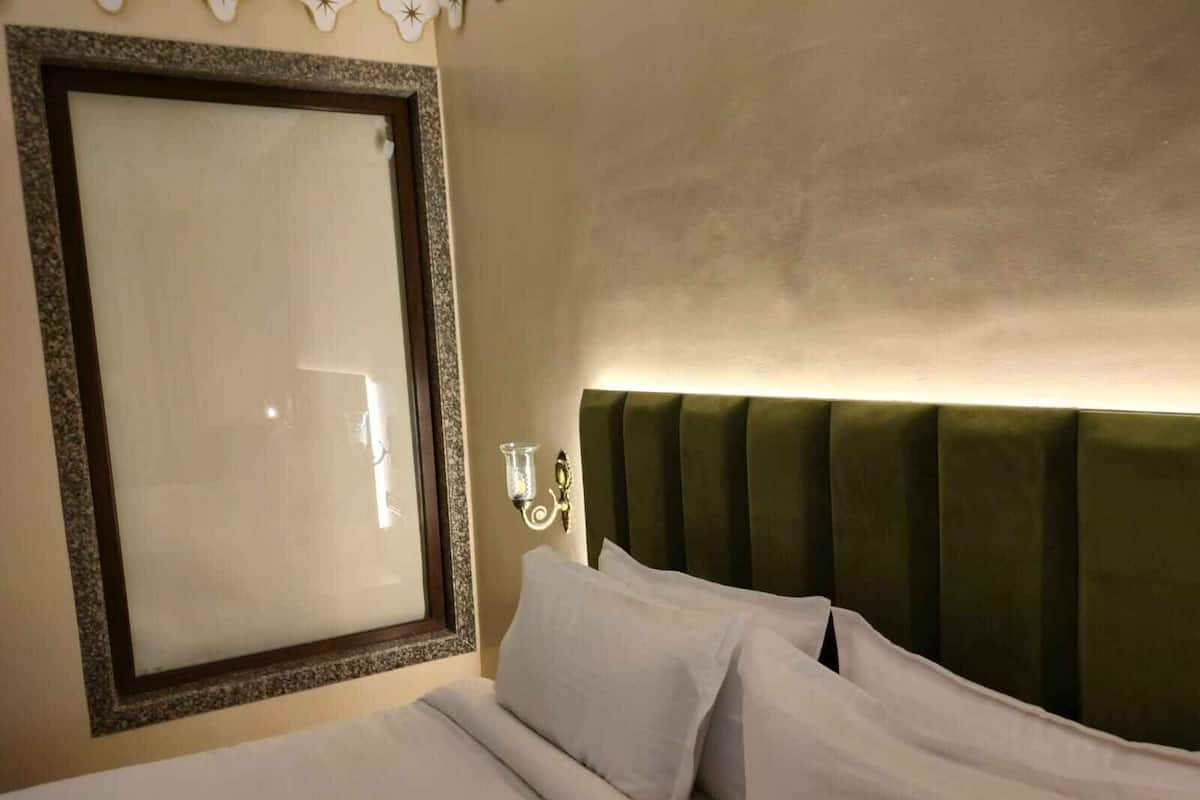 A hotel room with a large mirror, a green upholstered headboard, and a bedside table with a lamp.