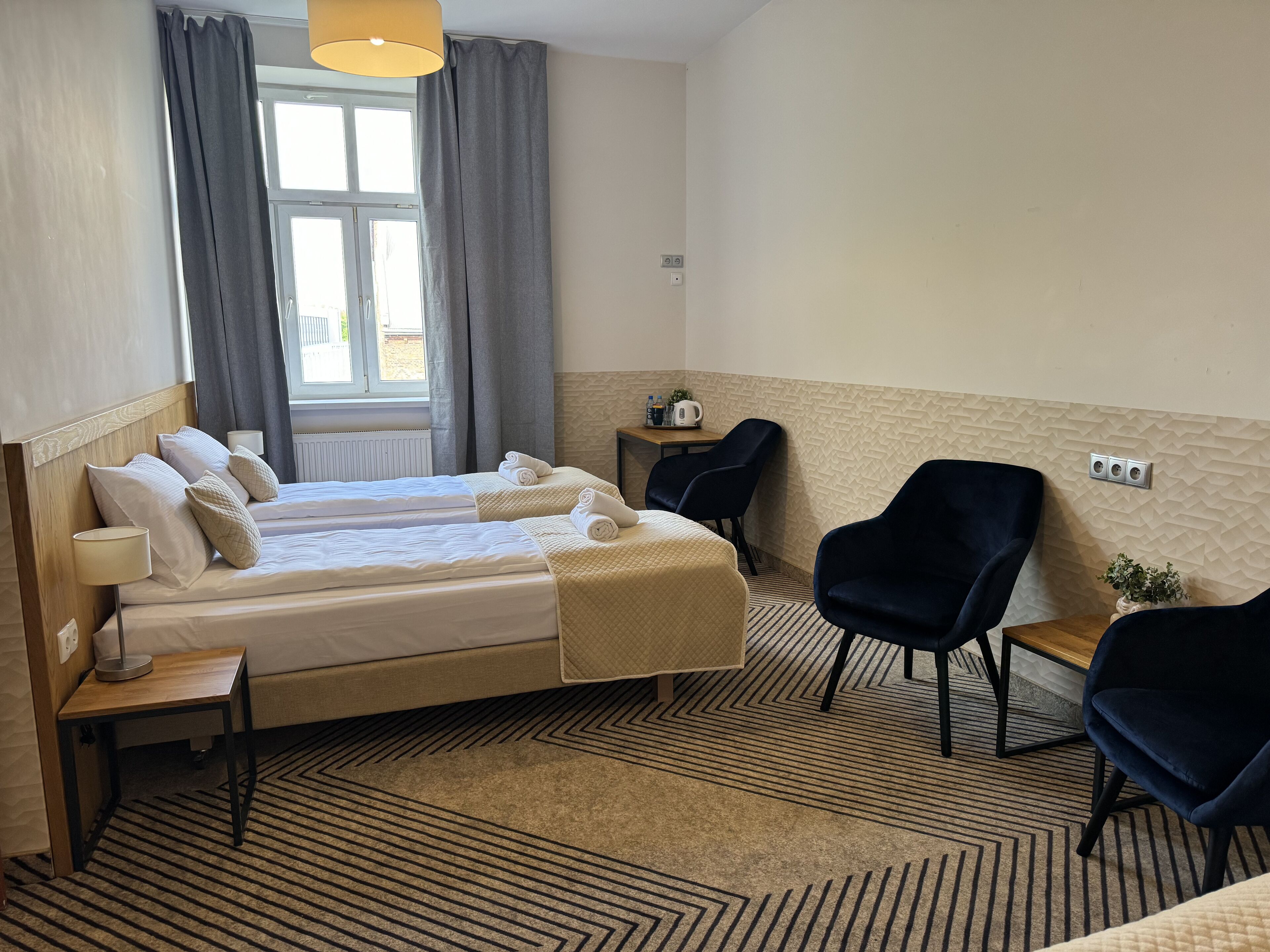 A hotel room with two beds, a small table, two chairs, a window with curtains, and a wall-mounted light fixture.