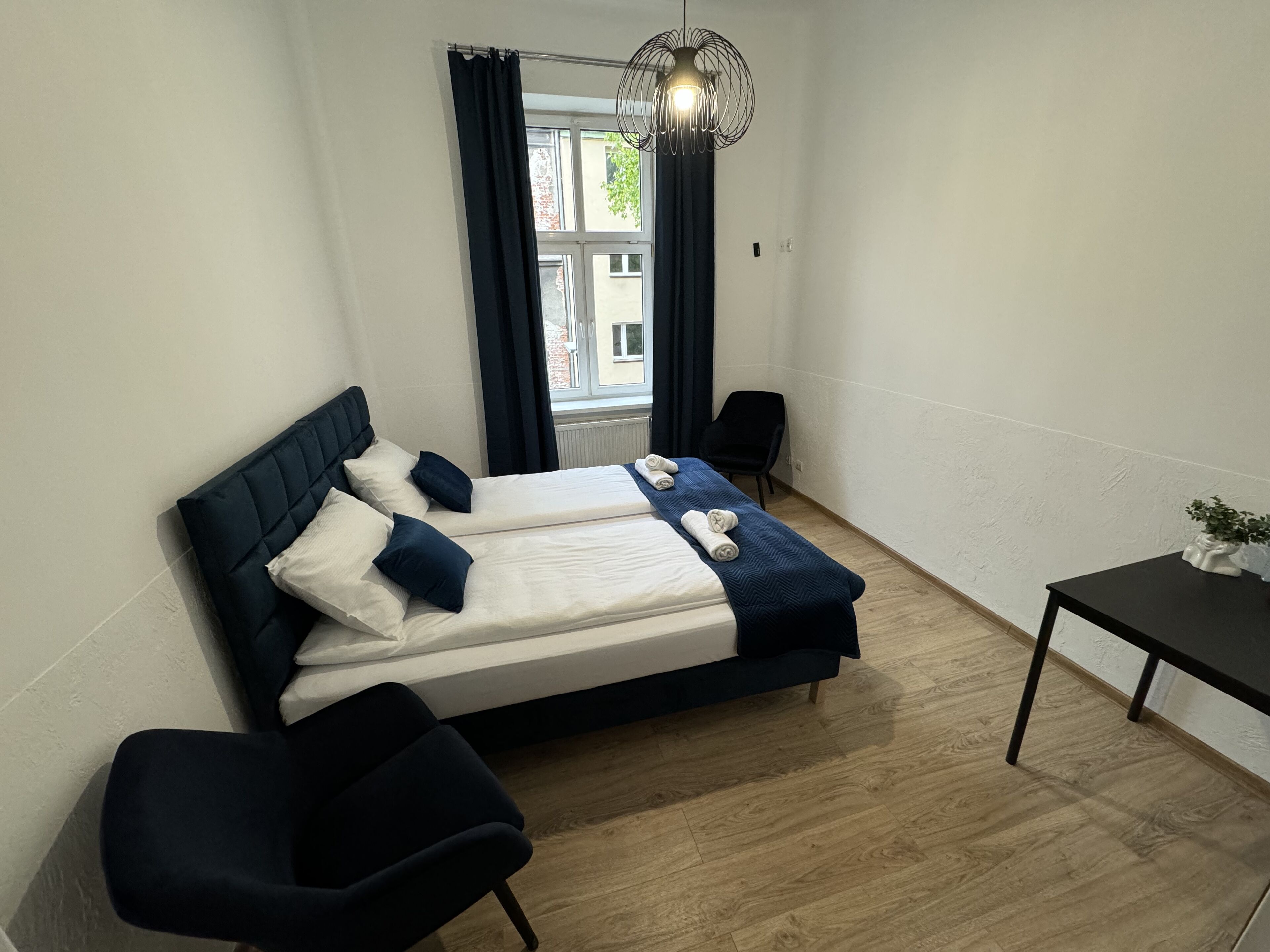 Un dormitorio pequeño y ordenado con cama, dos sillones, una mesita y una ventana con cortinas.