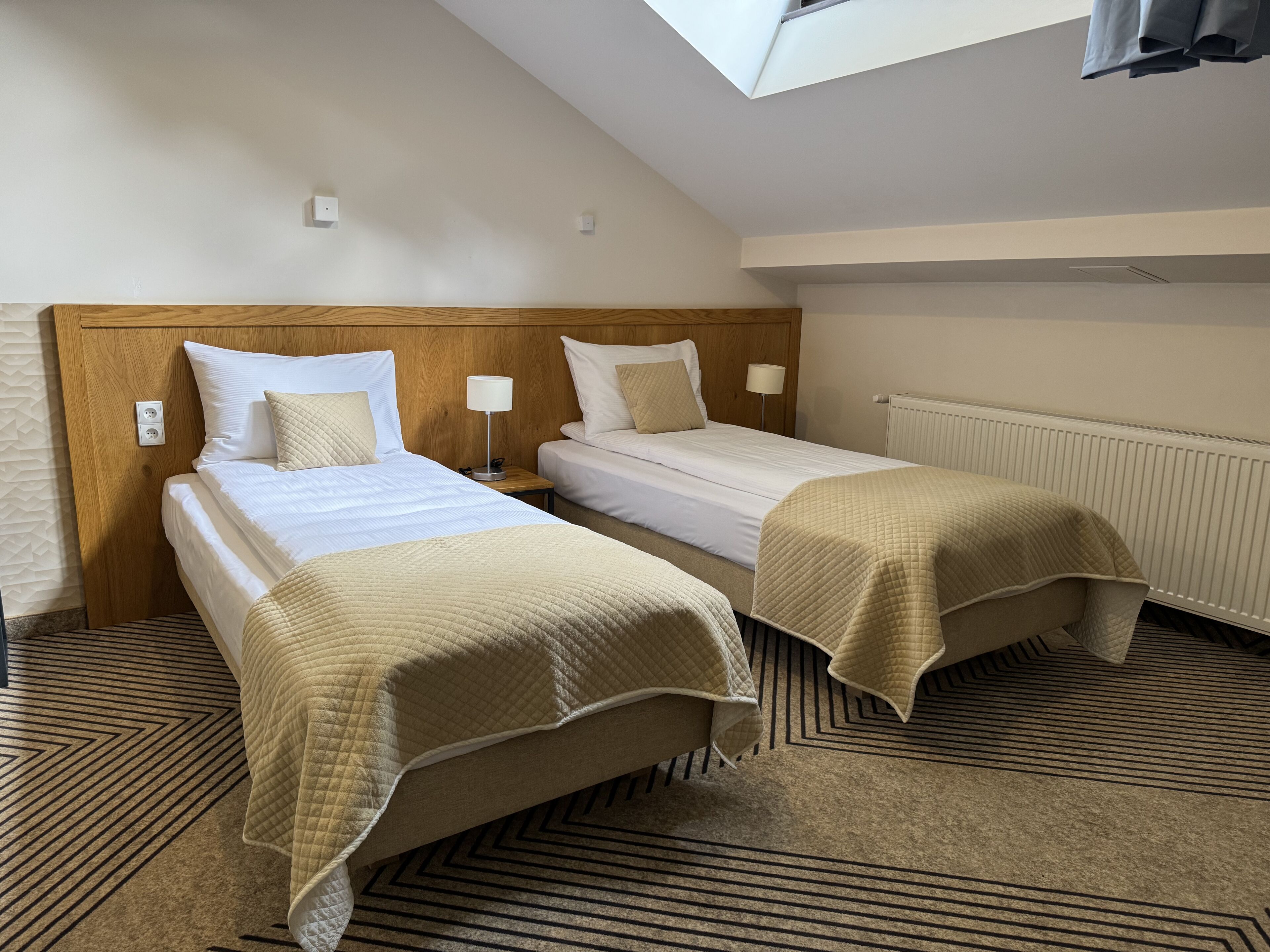 Habitación de hotel con dos camas individuales, cabecero de madera, tragaluz y radiador.