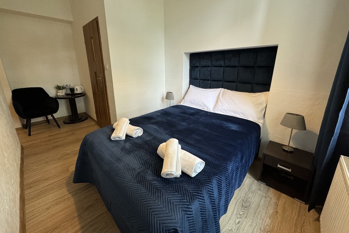 A hotel room with a bed, two towels, a chair, a small table, a lamp, and a nightstand.