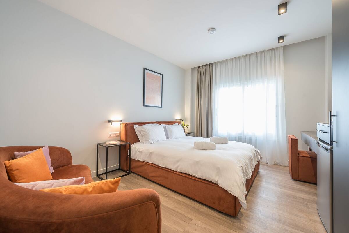 A modern hotel room with a large bed, a sofa, a desk, and a window with curtains.