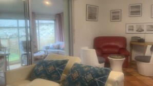 Un salón con sofá blanco, cojines estampados, un sillón rojo y una mesita auxiliar blanca. Se observan cuadros enmarcados en la pared y una puerta corrediza de cristal que da a un espacio exterior.