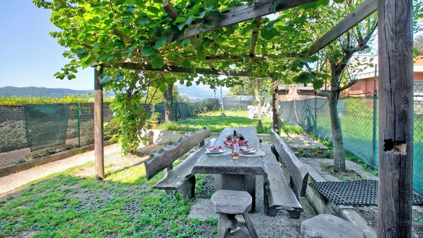 Conjunto de mesa e cadeiras de piquenique de madeira montado sob uma pérgula, com vista para um jardim cercado e colinas ao longe.
