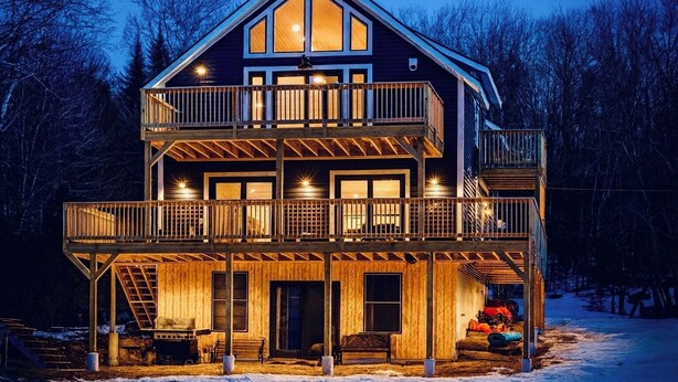 A multi-story wooden cabin with a balcony on the second floor, a covered porch, and a staircase leading to the upper levels. - Experience Lakeside Living with a Perfect Blend of Rustic Charm & Modern Luxury (Lunenburg)