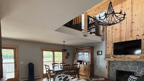 A spacious living room with a large wooden floor, a black sofa, and a stone fireplace. There is a dining area with wooden chairs and a table, and a kitchen with a stone backsplash. - Experience Lakeside Living with a Perfect Blend of Rustic Charm & Modern Luxury (Lunenburg)