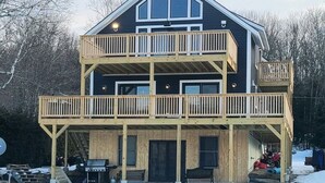 A two-story house with a balcony on the upper floor, a covered front porch, and a stone foundation. - Experience Lakeside Living with a Perfect Blend of Rustic Charm & Modern Luxury (Lunenburg)