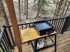 A portable grill on a wooden deck with a railing, surrounded by trees.