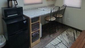 A compact kitchen with a microwave, coffee maker, and a small refrigerator. There are two windows with blinds, a wooden chair, and a small table. - Tiny Treehouse Cabin | Bluff Woodlands #1 (Elba)