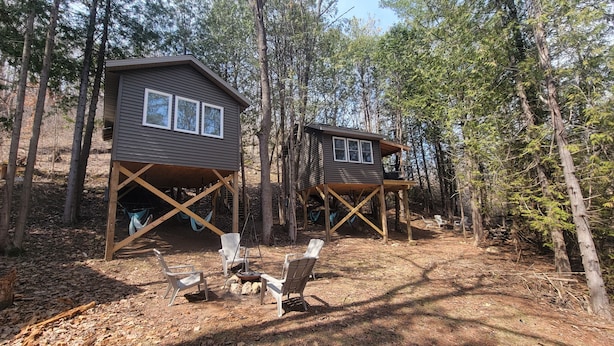 Two wooden cabins elevated on stilts in a forest, with outdoor seating and hammocks. - Tiny Treehouse Cabin | Bluff Woodlands #1 (Elba)