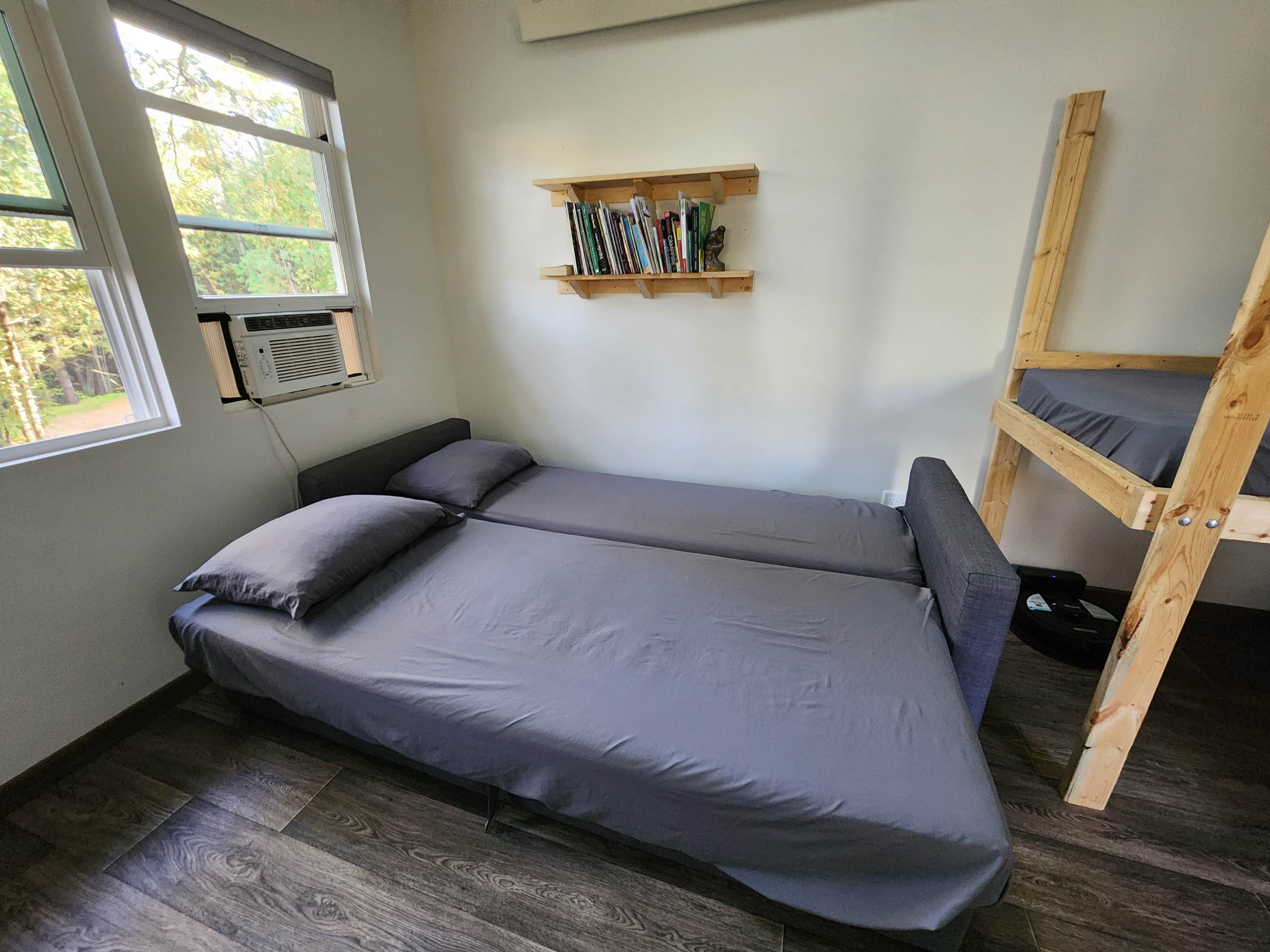Une petite chambre avec un lit simple, une fenêtre, une étagère avec des livres et un lit superposé.