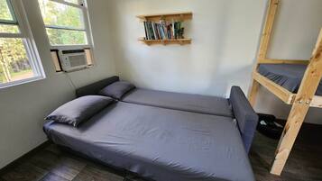 Une petite chambre avec un lit simple, une fenêtre, une étagère avec des livres et un lit superposé.